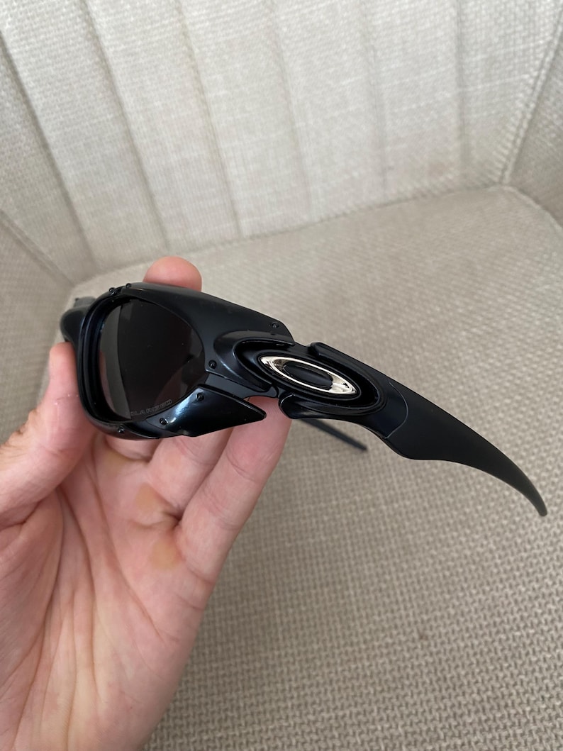 May include: Black sunglasses with a sleek, modern design. The sunglasses feature dark lenses and a curved frame with a metallic accent. The arms of the sunglasses are long and slightly curved.