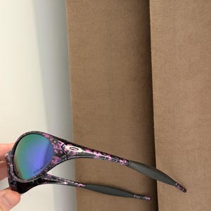 May include: Black and pink floral patterned sunglasses with mirrored lenses. The sunglasses have a black frame with pink floral accents and gray ear pieces. The lenses have a blue-green tint.