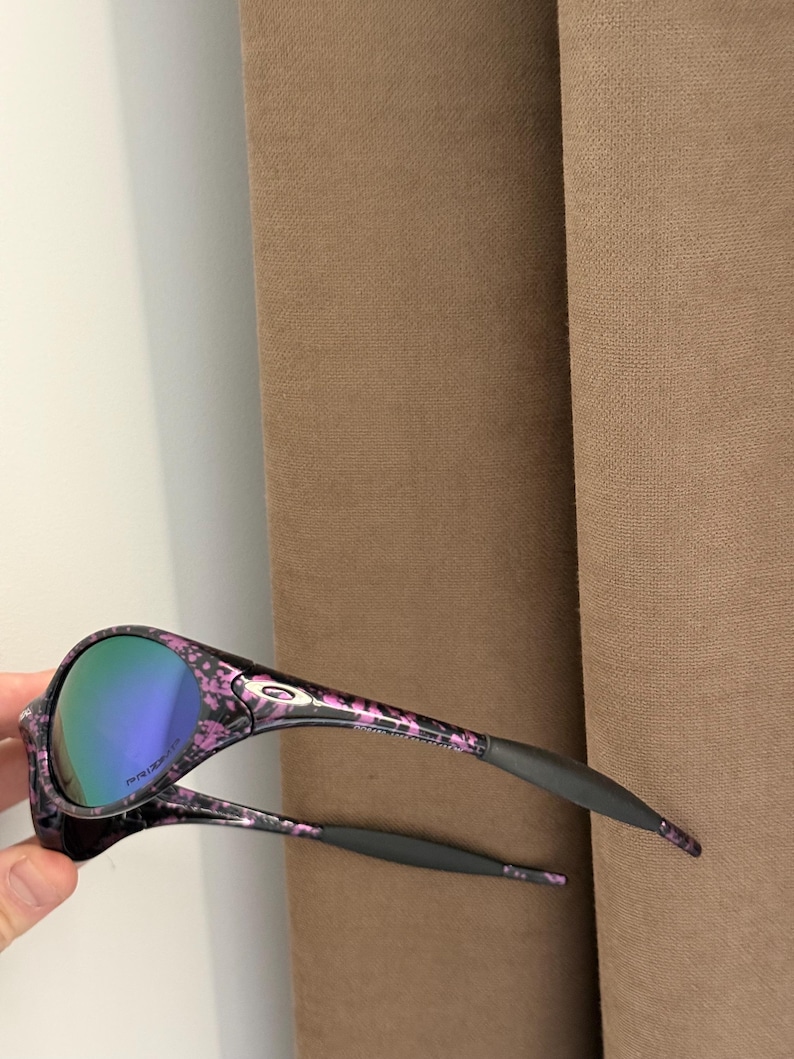May include: Black and pink floral patterned sunglasses with mirrored lenses. The sunglasses have a black frame with pink floral accents and gray ear pieces. The lenses have a blue-green tint.