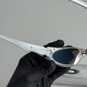 May include: Silver-framed sunglasses with blue lenses. The sleek, modern design features a metallic finish and curved temples. The sunglasses are held by a gloved hand, suggesting careful handling. The overall aesthetic is futuristic and stylish.