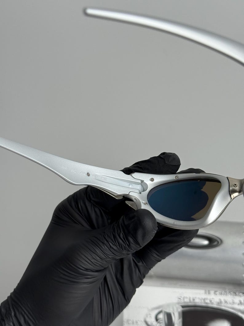 May include: Silver-framed sunglasses with blue lenses. The sleek, modern design features a metallic finish and curved temples. The sunglasses are held by a gloved hand, suggesting careful handling. The overall aesthetic is futuristic and stylish.