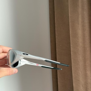 May include: Silver sunglasses with a dark lens. The sunglasses feature a modern design with black temple tips and a small red and white emblem. The lenses are mirrored, reflecting the environment. The frame is metallic.