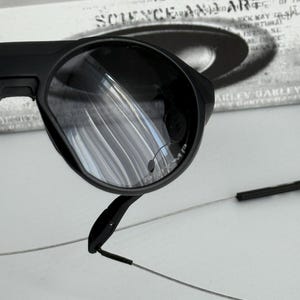 K&ouml;nnte beinhalten: Nahaufnahme einer schwarzen Sonnenbrille mit runder Linsenform. Die Brille hat ein schlankes, modernes Design mit d&uuml;nnen, silberfarbenen B&uuml;geln. Die Gl&auml;ser reflektieren die Umgebung. Der Markenname ist auf dem Glas sichtbar.