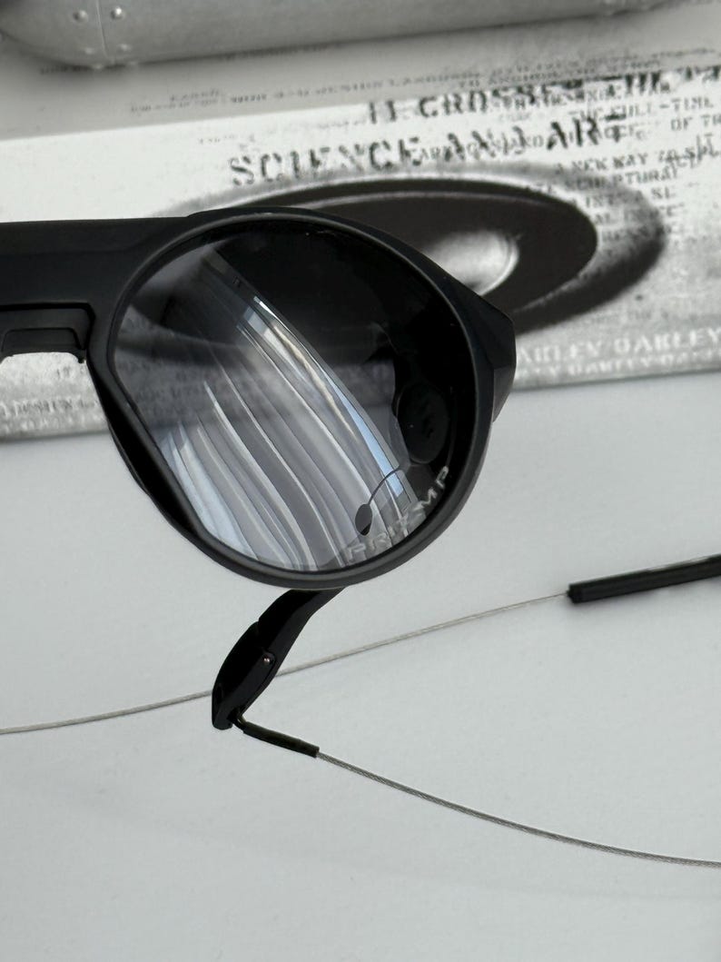 K&ouml;nnte beinhalten: Nahaufnahme einer schwarzen Sonnenbrille mit runder Linsenform. Die Brille hat ein schlankes, modernes Design mit d&uuml;nnen, silberfarbenen B&uuml;geln. Die Gl&auml;ser reflektieren die Umgebung. Der Markenname ist auf dem Glas sichtbar.
