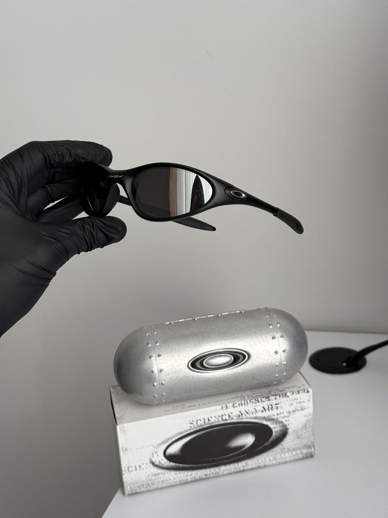 K&ouml;nnte beinhalten: Schwarze Sonnenbrille mit geschwungenem Rahmen und dunklen Gl&auml;sern. Die Sonnenbrille wird &uuml;ber einem silbernen Etui und einer Schachtel mit dem Text "SCIENCE AND ART" gehalten. Das Etui und die Schachtel tragen das Oakley-Logo.