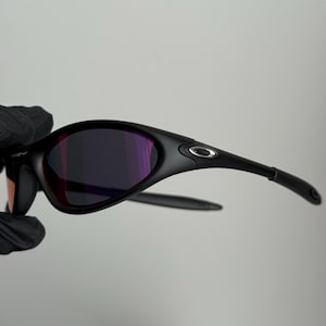 May include: Black sunglasses with a curved frame and purple-tinted lenses. The sunglasses have a sleek, modern design with a small silver logo on the temple. The lenses reflect a range of colors.