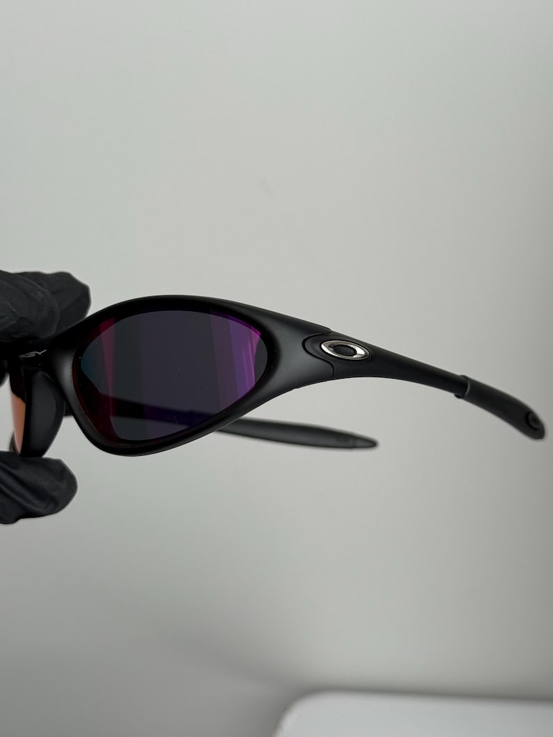 May include: Black sunglasses with a curved frame and purple-tinted lenses. The sunglasses have a sleek, modern design with a small silver logo on the temple. The lenses reflect a range of colors.