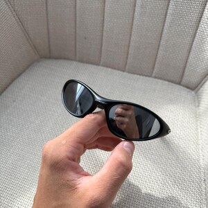 May include: Black oval sunglasses with dark lenses. The sunglasses have a sleek, modern design. The frame is a glossy black. The sunglasses are held in a hand.