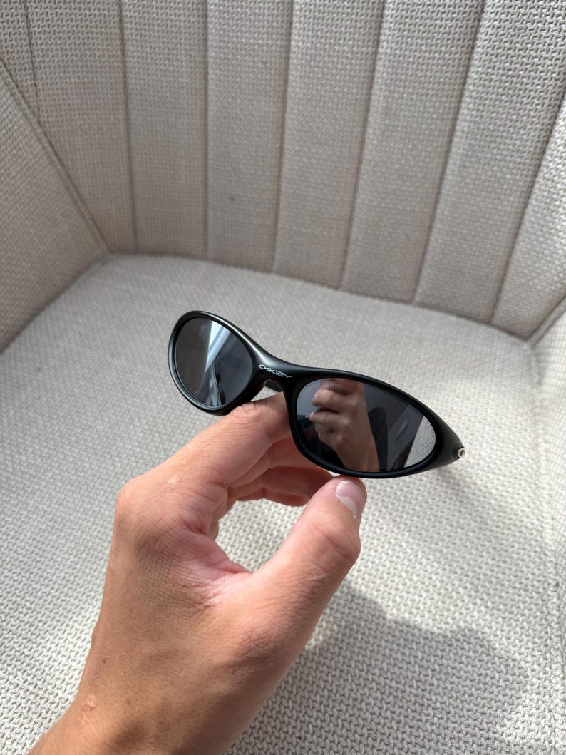 May include: Black oval sunglasses with dark lenses. The sunglasses have a sleek, modern design. The frame is a glossy black. The sunglasses are held in a hand.