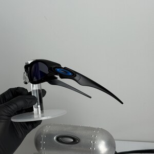 May include: Black and blue sunglasses with a sleek, modern design. The sunglasses have a black frame with blue accents and dark lenses. A silver metal case and a white box with text are also visible.