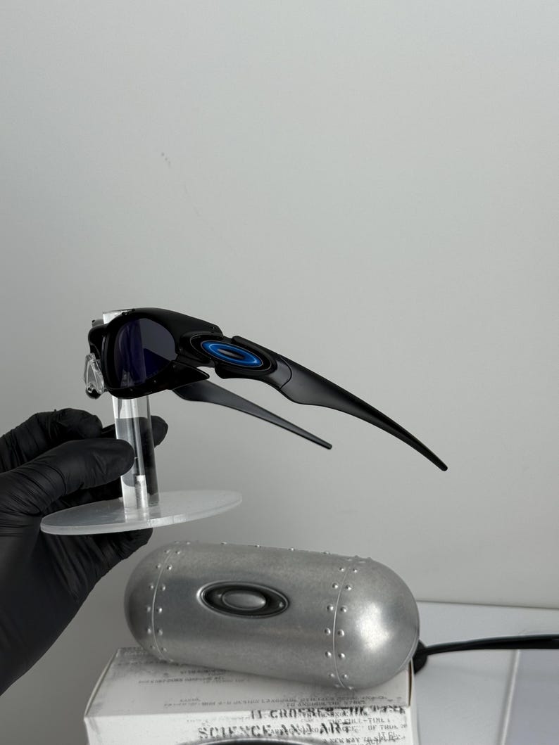 May include: Black and blue sunglasses with a sleek, modern design. The sunglasses have a black frame with blue accents and dark lenses. A silver metal case and a white box with text are also visible.