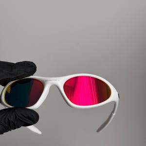 May include: White sunglasses with red-tinted lenses. The sunglasses have a modern design, with the brand name on the frame. The lenses reflect a bright, colourful hue.