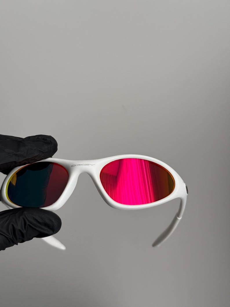 May include: White sunglasses with red-tinted lenses. The sunglasses have a modern design, with the brand name on the frame. The lenses reflect a bright, colourful hue.