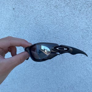 May include: Black Oakley sunglasses with a curved design and reflective lenses. The sunglasses have a sleek, modern aesthetic, with a dark frame and a subtle logo on the temple.