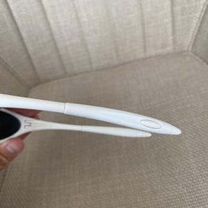 Puede incluir: Primer plano de gafas de sol blancas con lentes negras. Las gafas de sol tienen un dise&ntilde;o elegante y moderno con una montura y patillas finas. Las patillas son rectas y terminan en una punta curva.