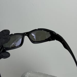 May include: Black sunglasses with a curved frame and dark lenses. The sunglasses have a sleek, modern design. The image also shows a silver case with a black oval logo.