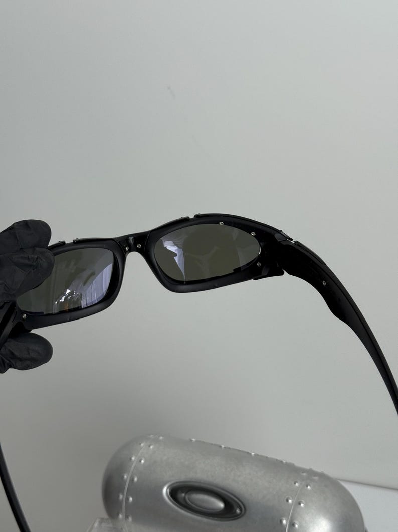 May include: Black sunglasses with a curved frame and dark lenses. The sunglasses have a sleek, modern design. The image also shows a silver case with a black oval logo.