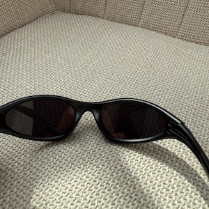 May include: Black sunglasses with a streamlined, curved shape. The lenses are dark, and the frames are matte black. The sunglasses are placed on a textured, light-coloured surface, implying a relaxed setting. The overall style is contemporary and sporty.