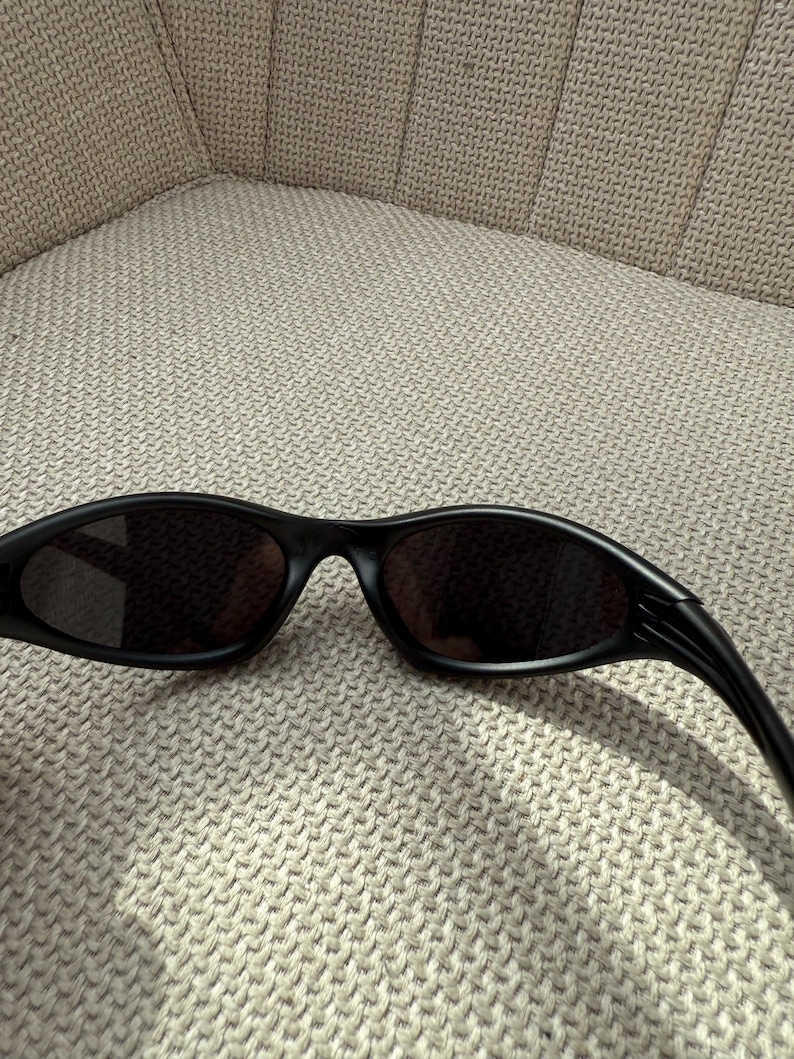 May include: Black sunglasses with a streamlined, curved shape. The lenses are dark, and the frames are matte black. The sunglasses are placed on a textured, light-coloured surface, implying a relaxed setting. The overall style is contemporary and sporty.