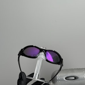 May include: Black sunglasses with purple lenses are displayed on a clear stand. The sunglasses have a sleek, modern design. The lenses reflect light, showing a vibrant purple hue. The frame is black and appears to be made of a durable material.