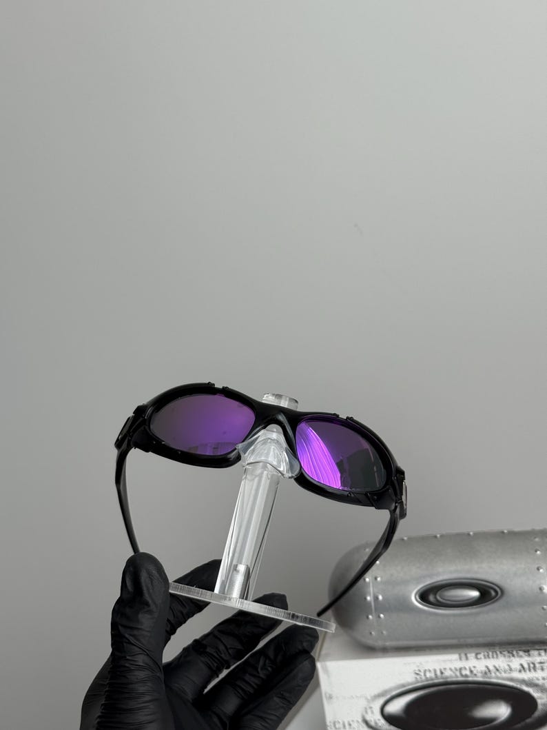 May include: Black sunglasses with purple lenses are displayed on a clear stand. The sunglasses have a sleek, modern design. The lenses reflect light, showing a vibrant purple hue. The frame is black and appears to be made of a durable material.