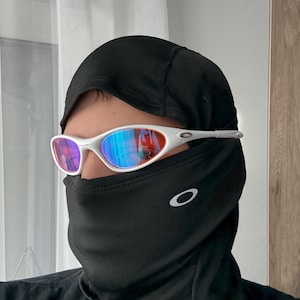 May include: White-framed sunglasses with a mirrored lens, worn with a black balaclava. The sunglasses have a sporty design, and the balaclava covers the lower face and neck. The Oakley logo is visible on the balaclava.