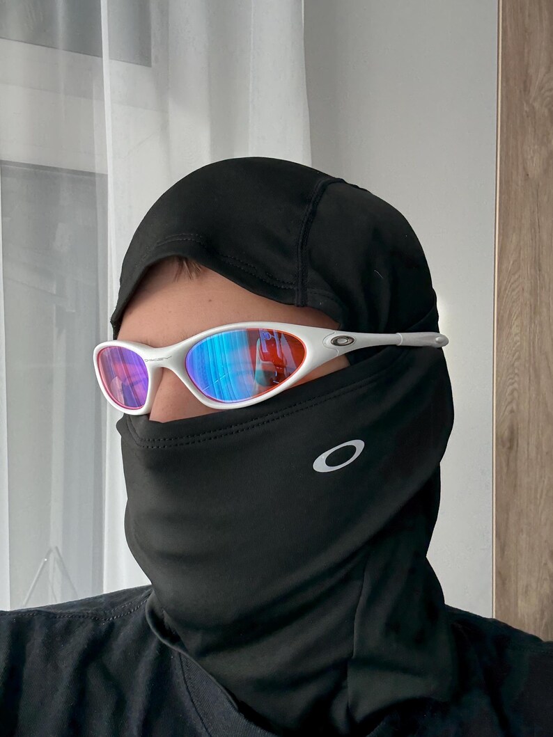 May include: White-framed sunglasses with a mirrored lens, worn with a black balaclava. The sunglasses have a sporty design, and the balaclava covers the lower face and neck. The Oakley logo is visible on the balaclava.