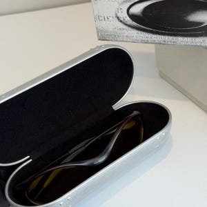 K&ouml;nnte beinhalten: Ge&ouml;ffnete silberne Sonnenbrillenetui mit einer Sonnenbrille darin. Die Sonnenbrille hat einen dunklen Rahmen und dunkle Gl&auml;ser. Das Etui ist ge&ouml;ffnet und zeigt ein schwarzes Innenfutter. Eine wei&szlig;e Schachtel mit dem Wort "SCIENCE" ist im Hintergrund.