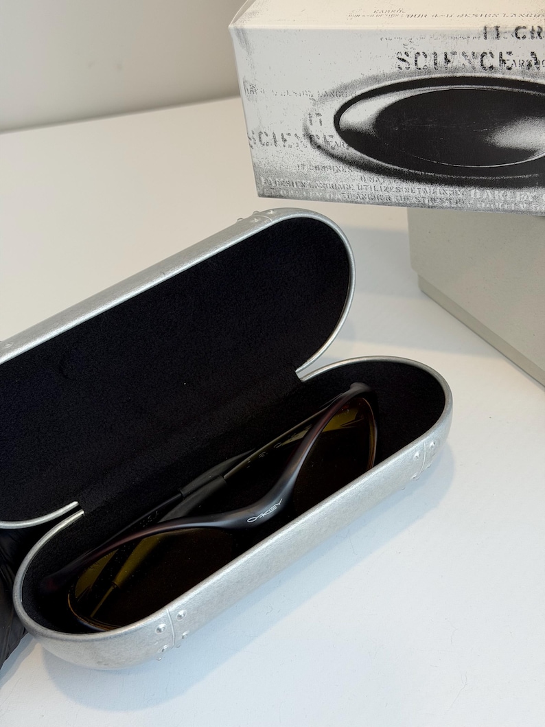K&ouml;nnte beinhalten: Ge&ouml;ffnete silberne Sonnenbrillenetui mit einer Sonnenbrille darin. Die Sonnenbrille hat einen dunklen Rahmen und dunkle Gl&auml;ser. Das Etui ist ge&ouml;ffnet und zeigt ein schwarzes Innenfutter. Eine wei&szlig;e Schachtel mit dem Wort "SCIENCE" ist im Hintergrund.