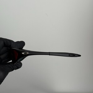 May include: Black sunglasses with a sleek, modern design. The frames are black, and the lenses appear to be a dark red or brown. The sunglasses are held by a gloved hand.
