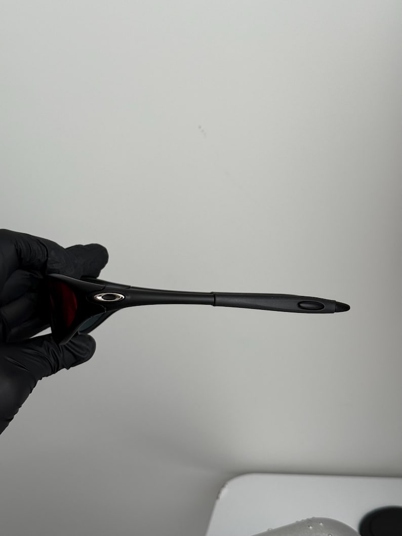 May include: Black sunglasses with a sleek, modern design. The frames are black, and the lenses appear to be a dark red or brown. The sunglasses are held by a gloved hand.