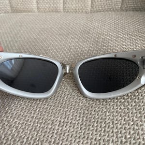 May include: Silver-framed sunglasses with dark lenses. The sunglasses have a sleek, modern design with a curved shape. The frames are a light silver color, and the lenses are black. The sunglasses are resting on a textured, beige surface.
