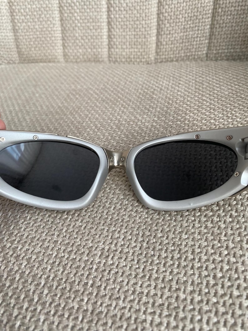 May include: Silver-framed sunglasses with dark lenses. The sunglasses have a sleek, modern design with a curved shape. The frames are a light silver color, and the lenses are black. The sunglasses are resting on a textured, beige surface.
