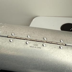 Puede incluir: Una funda de color plateado con un dise&ntilde;o remachado. La funda tiene bisagras y tiene el texto "CASE MADE IN CHINA" impreso en la superficie. La funda parece ser de metal.