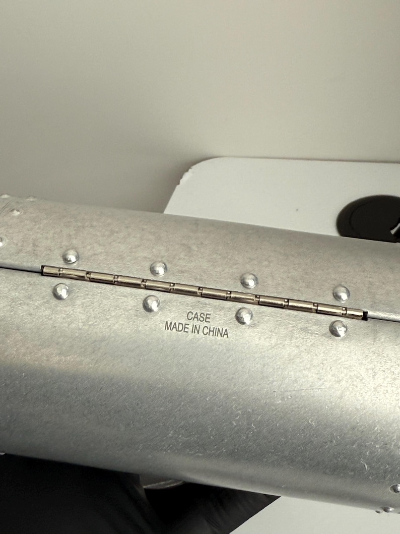 Puede incluir: Una funda de color plateado con un dise&ntilde;o remachado. La funda tiene bisagras y tiene el texto "CASE MADE IN CHINA" impreso en la superficie. La funda parece ser de metal.