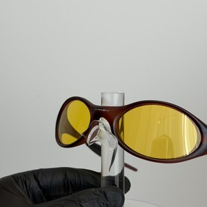 K&ouml;nnte beinhalten: Braune Sonnenbrille mit gelben Gl&auml;sern. Die Sonnenbrille hat einen dunkelbraunen Rahmen und wird von einem transparenten Displayst&auml;nder gehalten. Die Gl&auml;ser sind leuchtend gelb. Der Markenname "Oakley" ist auf dem Rahmen sichtbar.