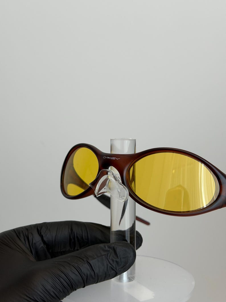 K&ouml;nnte beinhalten: Braune Sonnenbrille mit gelben Gl&auml;sern. Die Sonnenbrille hat einen dunkelbraunen Rahmen und wird von einem transparenten Displayst&auml;nder gehalten. Die Gl&auml;ser sind leuchtend gelb. Der Markenname "Oakley" ist auf dem Rahmen sichtbar.