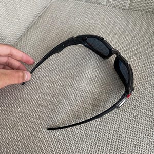 Op de afbeelding: Zwarte zonnebril met donkere glazen. De zonnebril heeft een strak, sportief design met een zwart montuur en pootjes. De glazen lijken donkergrijs of zwart van kleur te zijn.
