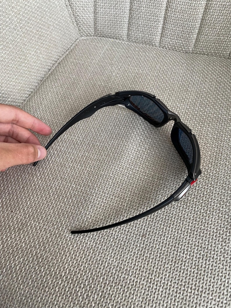 Op de afbeelding: Zwarte zonnebril met donkere glazen. De zonnebril heeft een strak, sportief design met een zwart montuur en pootjes. De glazen lijken donkergrijs of zwart van kleur te zijn.