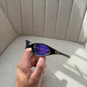 May include: Black sunglasses with purple-blue mirrored lenses. The sunglasses have a sleek, modern design with a black frame and arms. The lenses reflect a vibrant array of colours. The Oakley logo is visible on the arm.