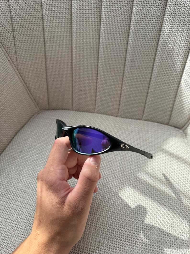 May include: Black sunglasses with purple-blue mirrored lenses. The sunglasses have a sleek, modern design with a black frame and arms. The lenses reflect a vibrant array of colours. The Oakley logo is visible on the arm.