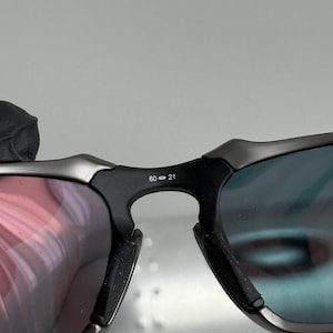May include: Close-up of sunglasses with a black frame and lenses that reflect red and blue hues. The bridge of the glasses has the numbers "60-21" printed on it. The sunglasses have a modern, sporty design.