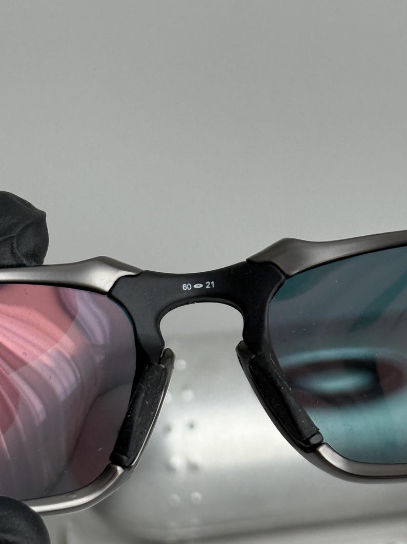 May include: Close-up of sunglasses with a black frame and lenses that reflect red and blue hues. The bridge of the glasses has the numbers "60-21" printed on it. The sunglasses have a modern, sporty design.