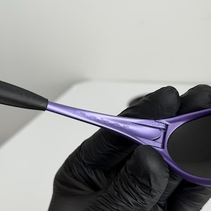 May include: Close-up of purple sunglasses with black lenses and arms. The sunglasses have the word "jacket" printed on the arm. The sunglasses are held by a person wearing black gloves.