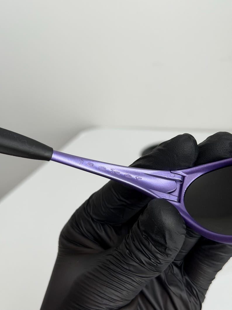 May include: Close-up of purple sunglasses with black lenses and arms. The sunglasses have the word "jacket" printed on the arm. The sunglasses are held by a person wearing black gloves.