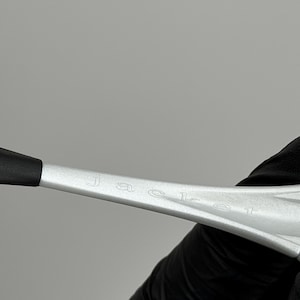 May include: Close-up of a silver object with the word "JACEL" engraved on it. The object has a black tip and is held by a person wearing a black glove. The background is a solid grey.