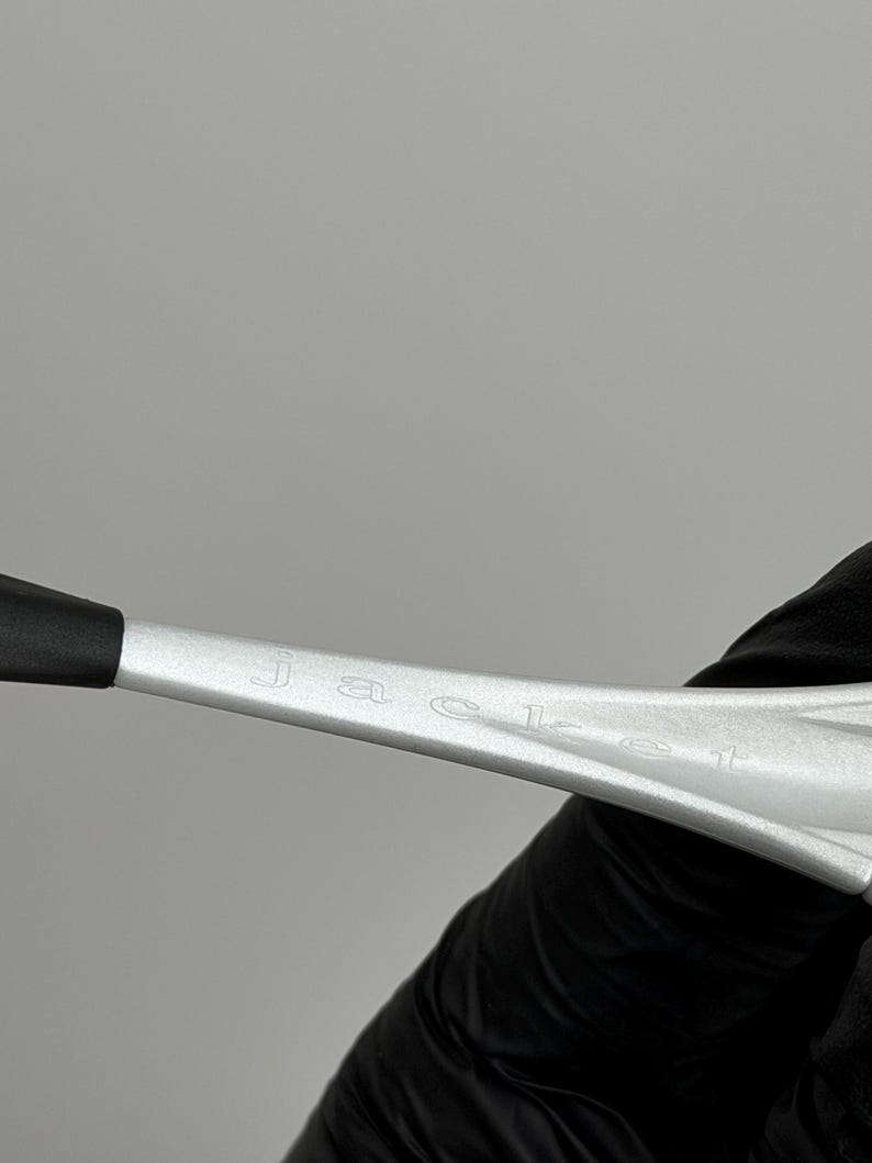 May include: Close-up of a silver object with the word "JACEL" engraved on it. The object has a black tip and is held by a person wearing a black glove. The background is a solid grey.