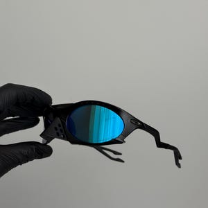 May include: Black sunglasses with blue mirrored lenses. The sunglasses have a sleek, modern design with a curved frame and unique temple arms. The lenses reflect a vibrant blue and green color.
