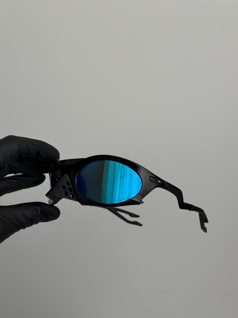 May include: Black sunglasses with blue mirrored lenses. The sunglasses have a sleek, modern design with a curved frame and unique temple arms. The lenses reflect a vibrant blue and green color.