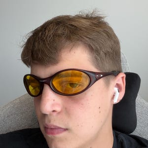 K&ouml;nnte beinhalten: Braune, ovale Sonnenbrille mit gelben Gl&auml;sern. Die Sonnenbrille hat einen dunkelbraunen Rahmen, und der Markenname "Oakley" ist oben auf dem Rahmen sichtbar. Die Gl&auml;ser reflektieren die Umgebung.